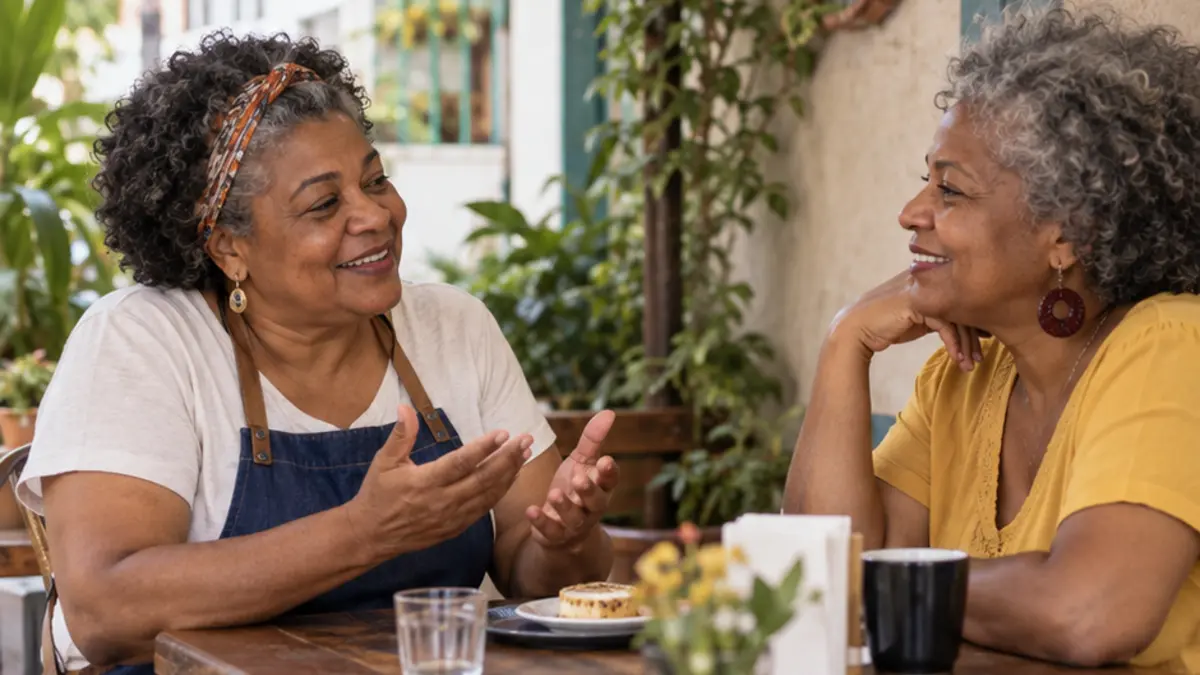 Duas mulheres conversam à mesa, uma de avental azul e outra de blusa amarela
