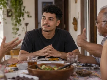 Jovem de camiseta preta está à mesa entre três adultos que gesticulam durante uma conversa